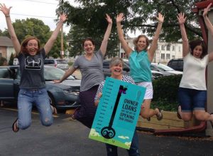 team of volunteers jumping for joy in a parking lot; person holding large successful fundraising tracker