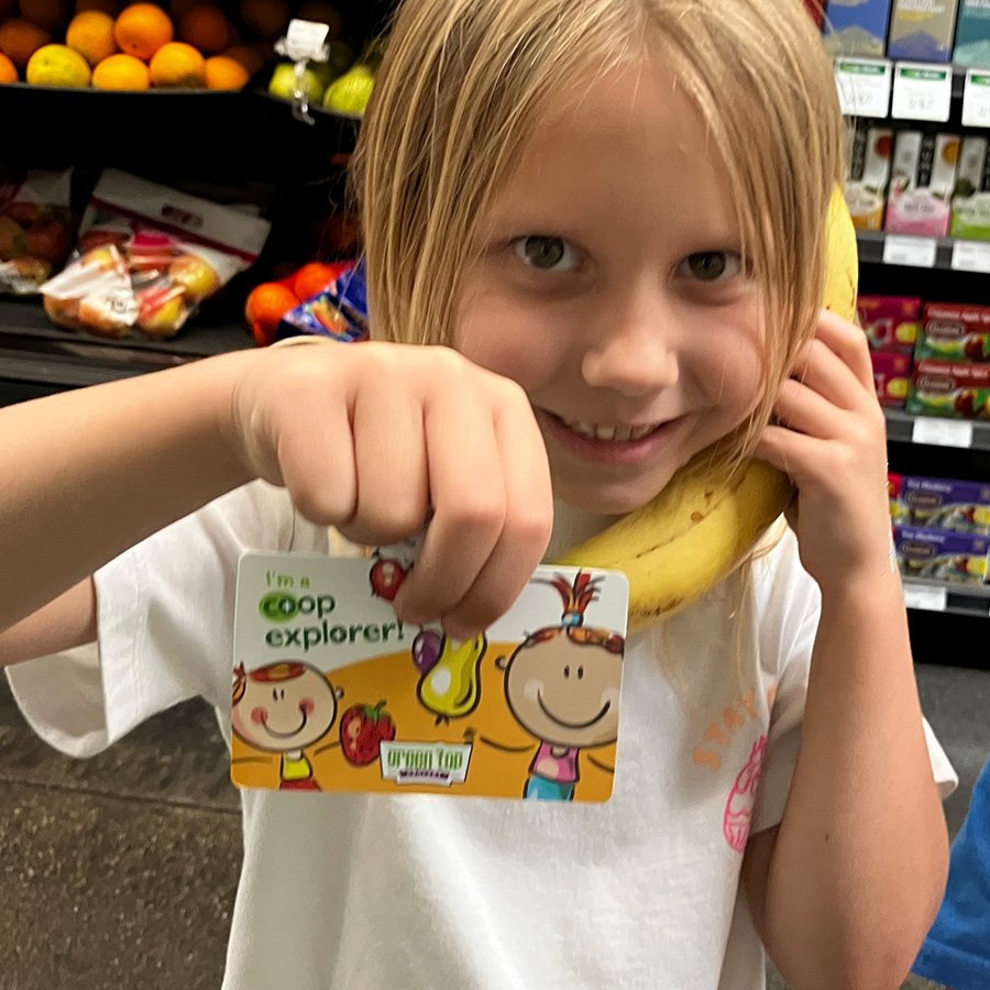 young girl holding a banana to her ear like a telephone, smiling at the camera and showing her co-op explorer card