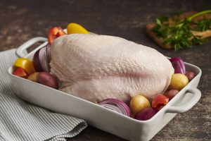 Uncooked bone-in turkey breast placed in a white ceramic roasting pan, surrounded by red and yellow potatoes, red onion wedges, and chunks of red and yellow bell peppers. The pan rests on a striped cloth atop a dark countertop, with a wooden cutting board and fresh parsley visible in the background. The scene suggests a rustic, home-style preparation for roasting, highlighting fresh ingredients and vibrant colors.