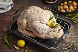 Raw, seasoned whole turkey resting in a black roasting pan with a metal rack, garnished with fresh dill and stuffed with lemon halves. Surrounding the pan are cooking ingredients and tools: whole lemon, garlic cloves, fresh herbs, red and yellow baby potatoes in a dish, kitchen scissors, and asparagus spears. The scene is set on a rustic kitchen counter, evoking a festive, home-cooked meal in preparation.