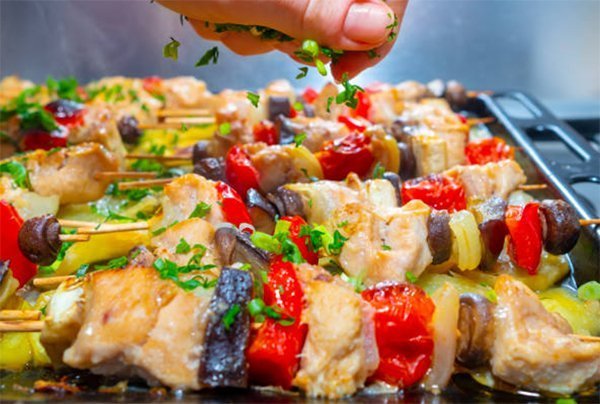 photo of a row of chicken and vegetable kabobs on a grill. A hand sprinkles fresh herbs from above.